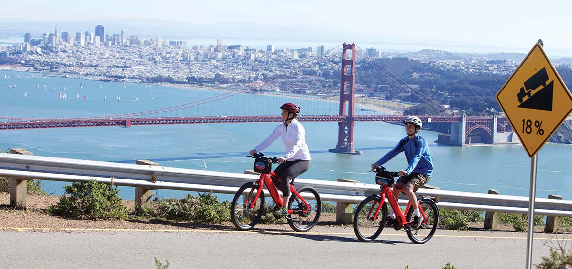 Two people riding tandem bike with San Francisco skyline in background